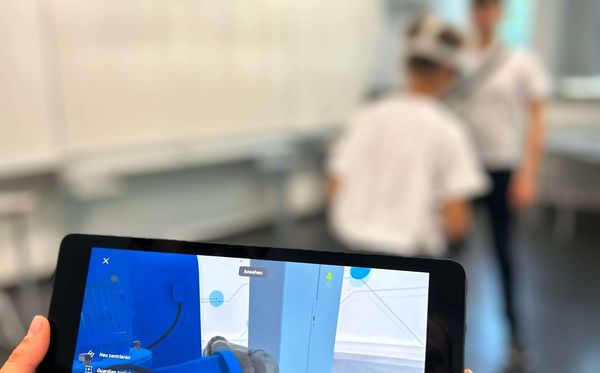 Zwei Schüler im Klassenzimmer spielen die VWE. Im Vordergrund ist ein Tablet abgebildet, welches das Bild aus der VR-Anwendung anzeigt.