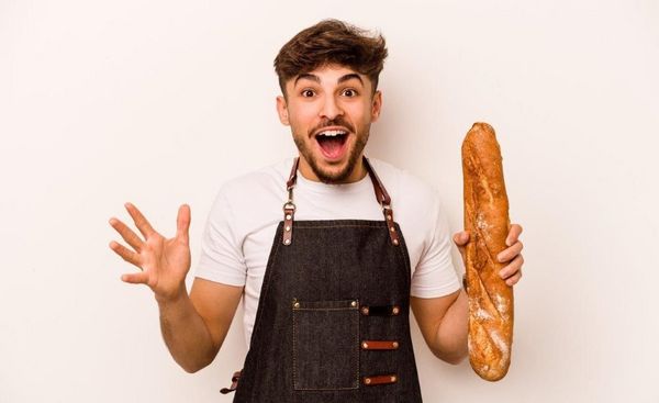 Junger Mann vor einem weißen Hintergrund. Er hält eine Baguettestange in seiner linken Hand und lächelt in die Kamera.